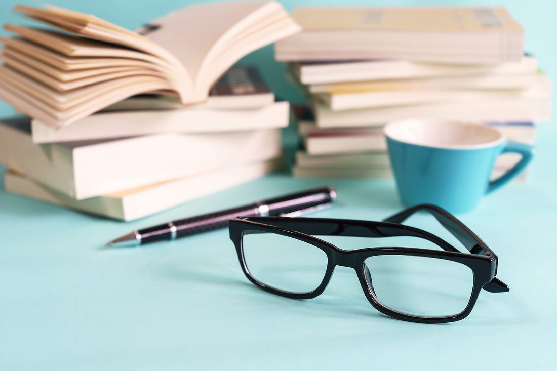 Stack of books with open book, black glasses and pen on turquoise surface next to a blue coffee cup, representing the Vibease vibrator experience with erotic audiobooks.