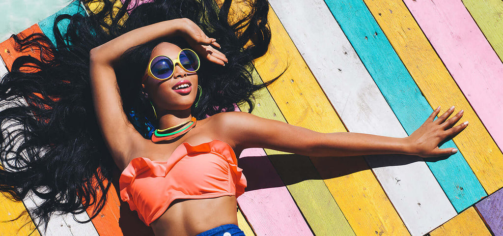 woman in orange bikini top and colorful sunglasses lying on rainbow-colored wooden floor promoting health benefits of masturbation