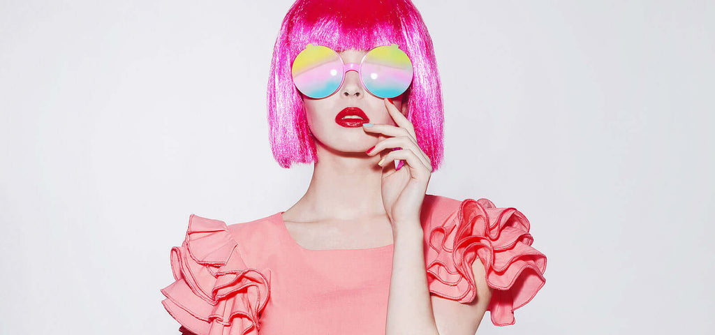 Woman with bright pink bob hairstyle and matching pink ruffled dress wearing reflective rainbow sunglasses and red lipstick touching her face