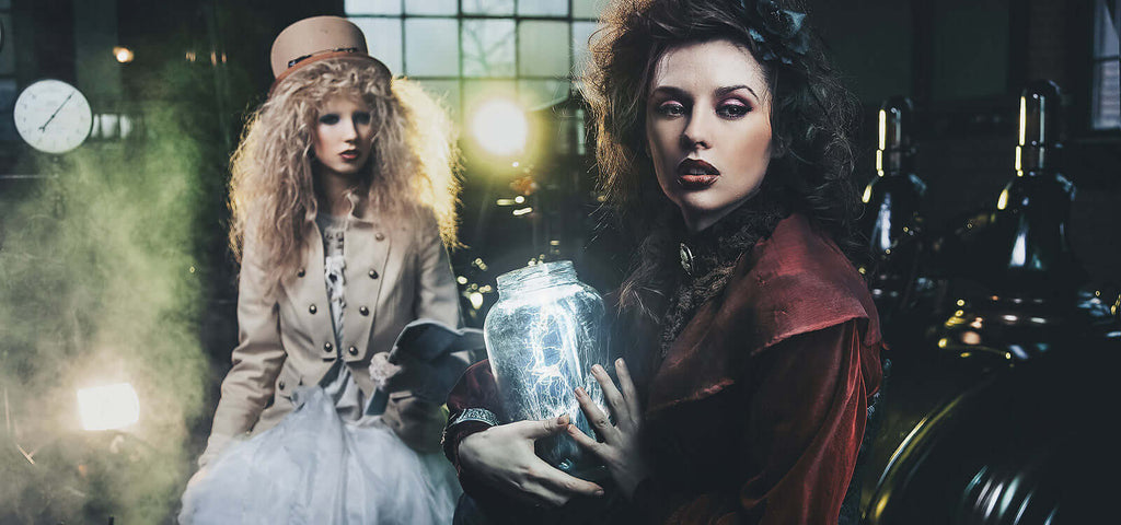 Two women in Victorian-style clothing in a dimly lit laboratory setting with one holding a glowing jar, evoking a mysterious and experimental "Sex Lab" atmosphere