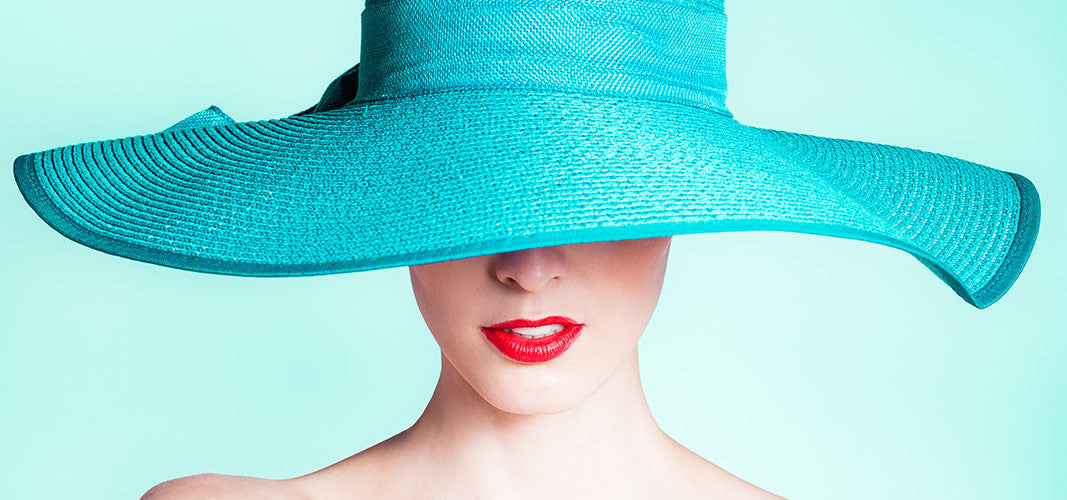 Woman wearing large turquoise hat covering eyes with red lipstick against turquoise background