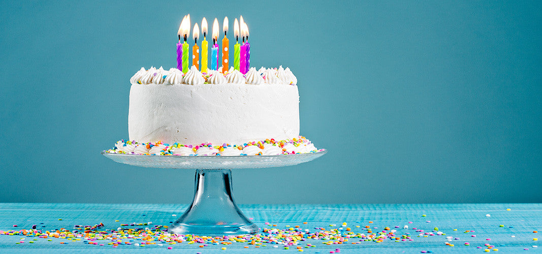White birthday cake with colorful lit candles and sprinkles on a glass cake stand celebrating Penchant's First Birthday